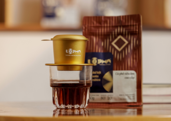 Gold LOPHA phin filter resting on a glass of black coffee, with a LOPHA coffee bag in the background